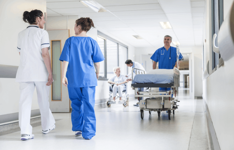 Drukke hal in een ziekenhuis met twee verpleegsters, een man met een rollend bed en een oudere vrouw in een rolstoel rondlopen.